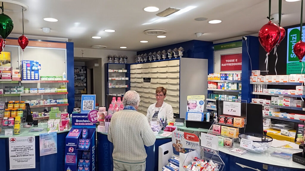 Interno di una farmacia con una cliente al banco servita da una farmacista, tra scaffali di prodotti e cassettiere dei farmaci