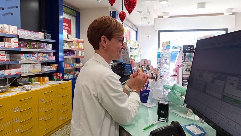Una farmacista in camice al banco di una farmacia, accanto al computer e alle cassettiere dei farmaci