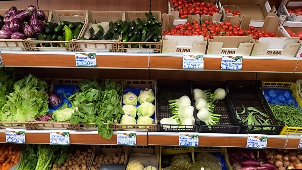 Scaffali di un negozio di ortofrutta con melanzane, zucchine, pomodorini, insalate, cavolfiori, finocchi e fagiolini nelle cassette con i cartellini dei prezzi