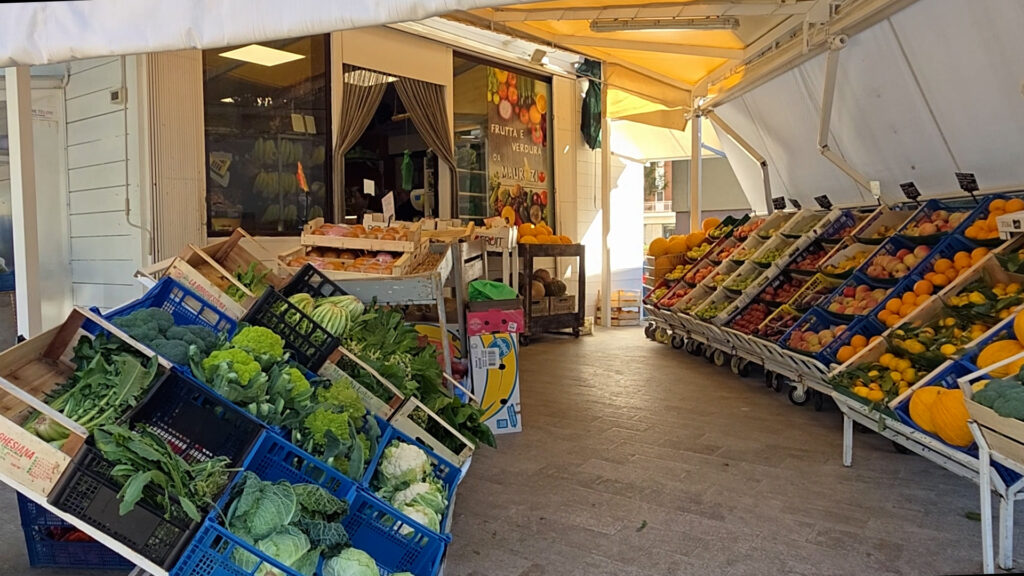 Cassette di cavoli, broccoli, cavolfiori e verdure a foglia e banchi di agrumi e frutta esposti sotto la tenda di un negozio di ortofrutta
